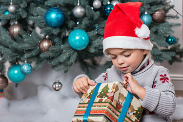Young girl wearing Santa hat, sits on floor in front of Christmas tree, opening present